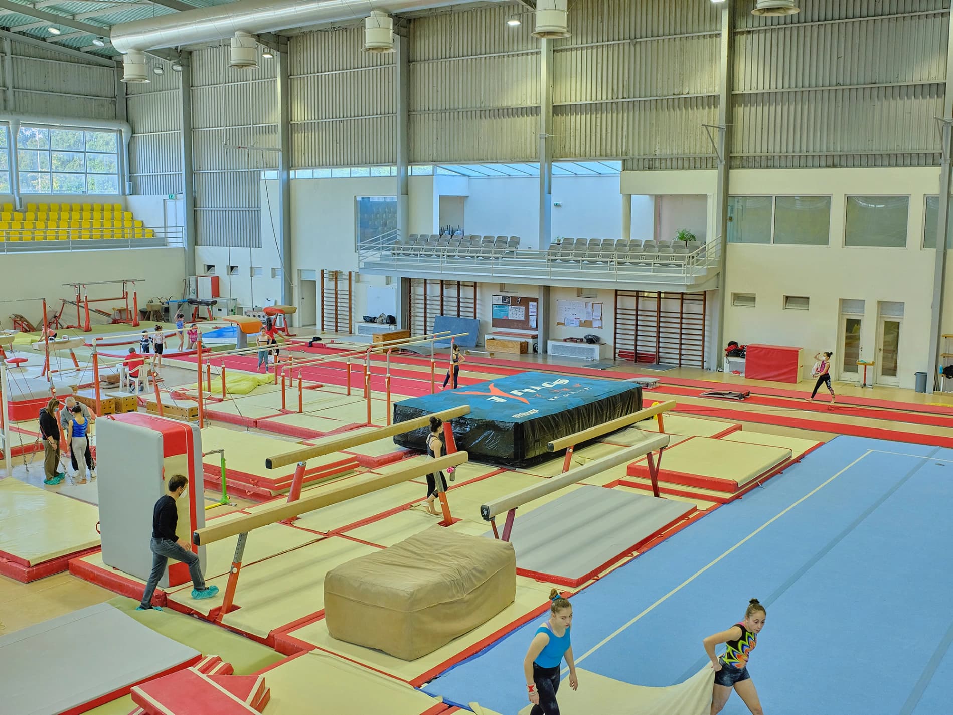 Indoor gymnastics hall with balance beams and red-bordered mats in the foreground and two gymnasts carrying a beige mat on the blue floor.