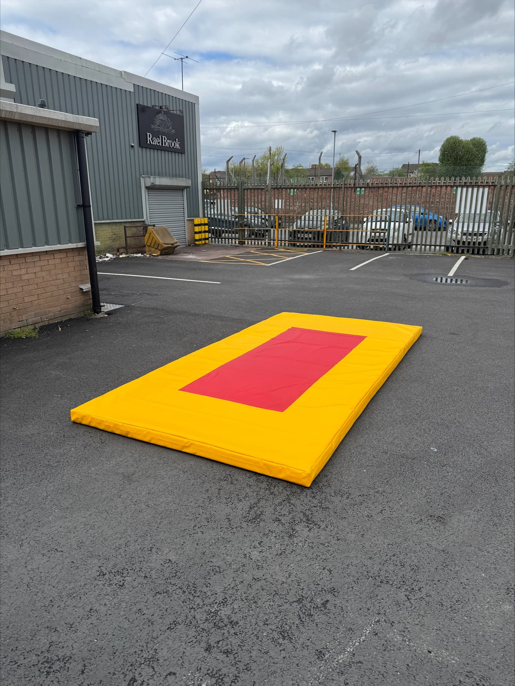 Bright yellow gymnastics training mat with a central red target rectangle on asphalt in a parking lot; building sign reads Rael Brook.
