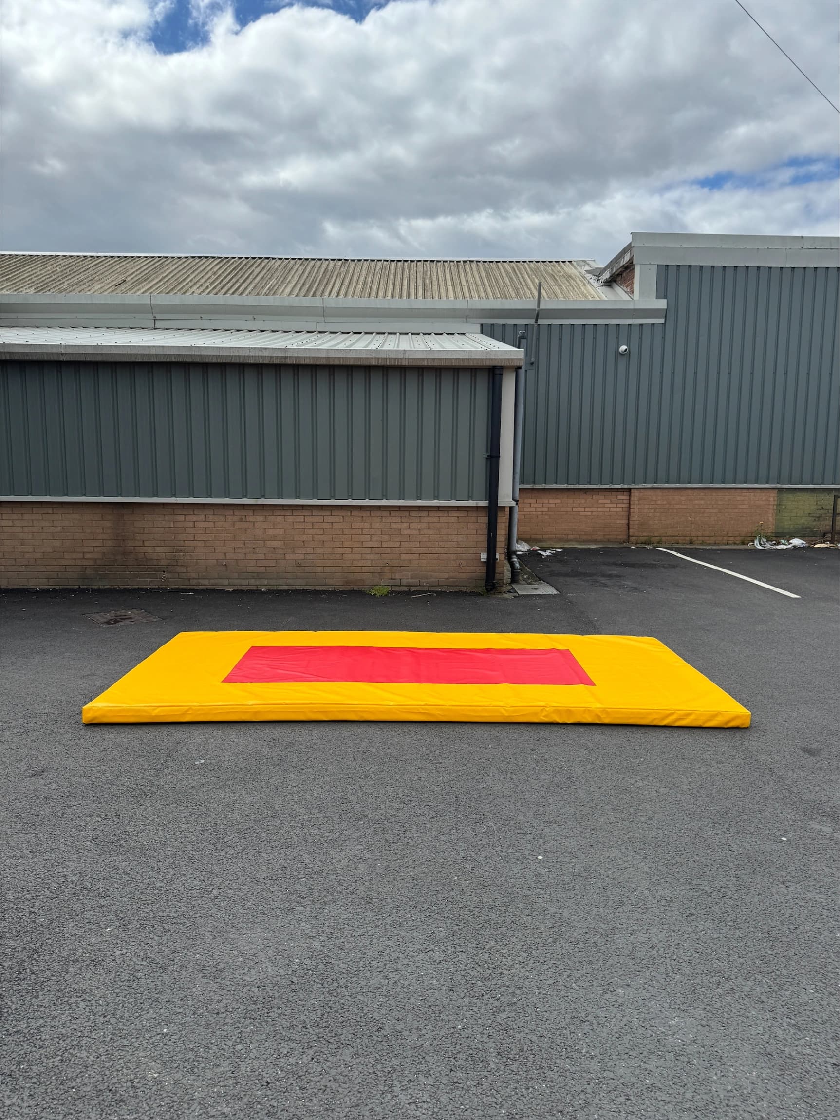 Bright yellow gymnastics mat with a central red target zone lying on asphalt outside a gray industrial building under cloudy sky.