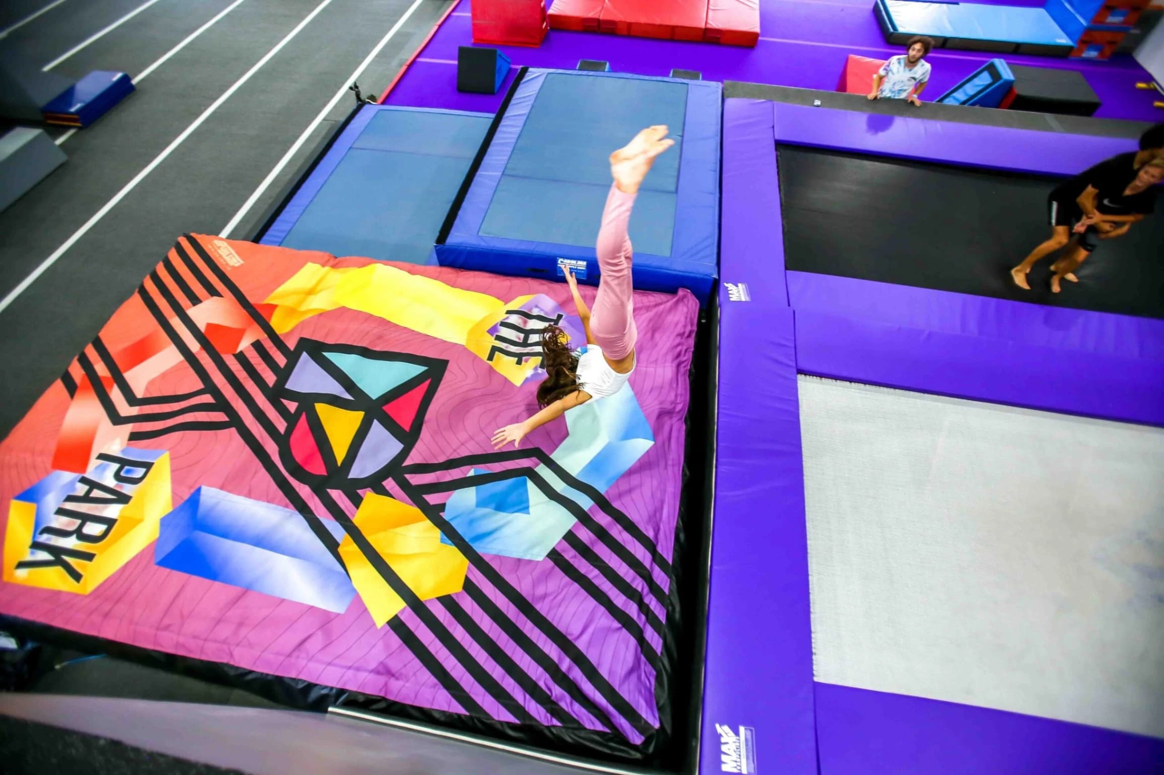 Girl flipping headfirst onto colorful airbag printed with geometric shapes and the word PARK, next to purple trampolines.